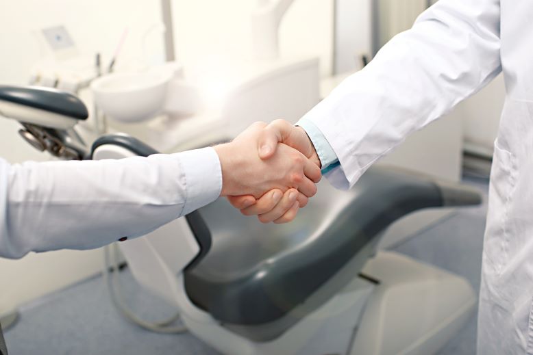 A man shaking hands with his dentist in front of a dental exam chair.